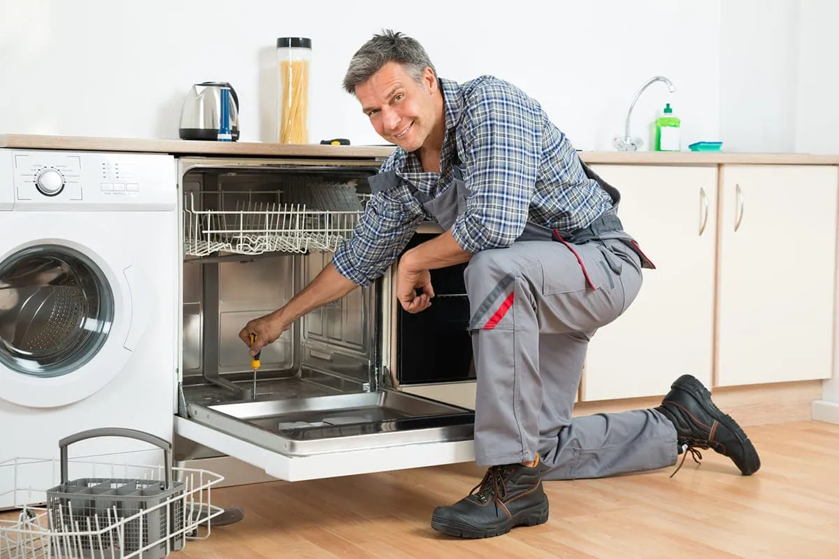 Técnico reparando un lavavajillas en una cocina, con el zócalo retirado para acceder a las bombas